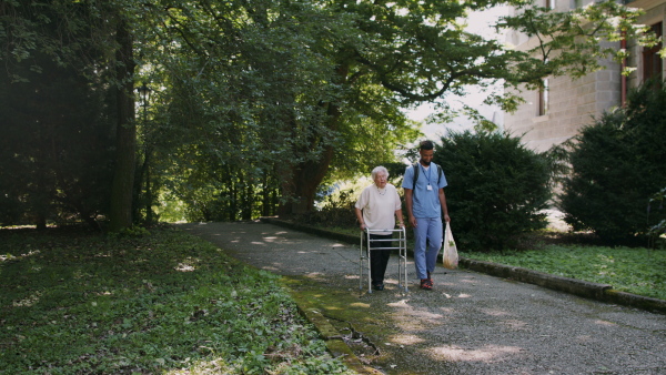 A senior woman and caregiver outdoors on a walk with walker in park, talking.