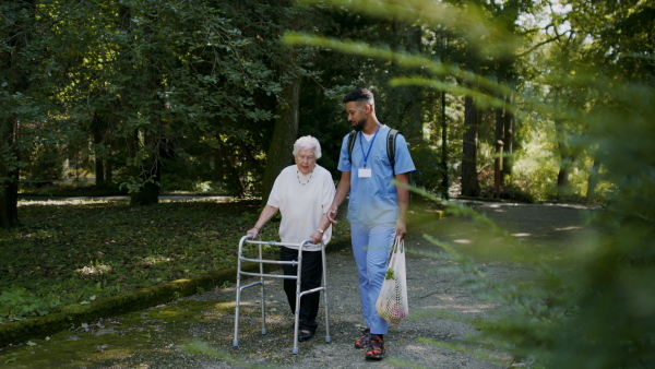 A senior woman and caregiver outdoors on a walk with walker in park, talking.