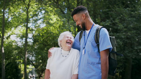 A senior woman and caregiver outdoors on a walk in park, talking and laughing.