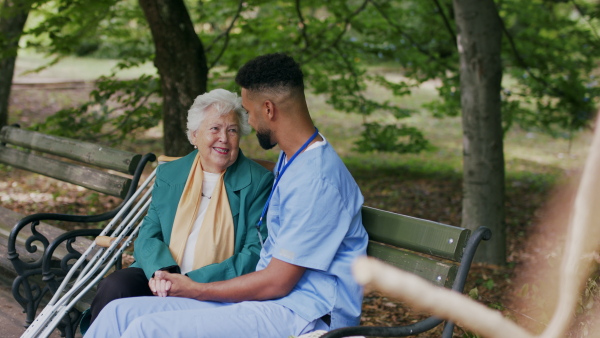 A caregiver man talking and supporting senior woman when sitting outdoors on bench in park.