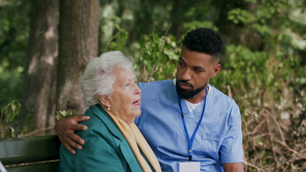 A caregiver man talking and supporting senior woman when sitting outdoors on bench in park.