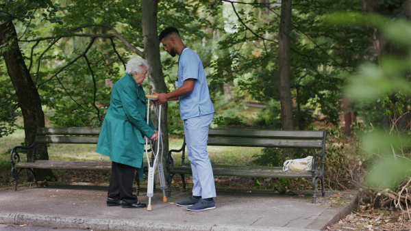 A caregiver man helping senior woman with crutches to sit on bench during walk outdoors in park.