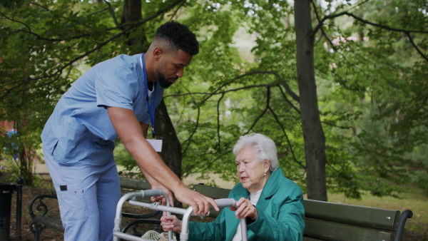 A caregiver man helping senior woman with walker to stand up from bench outdoors in park.