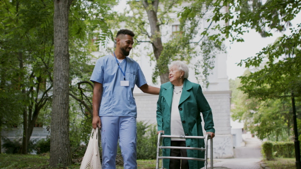 A senior woman and caregiver outdoors on a walk with walker in park, talking.