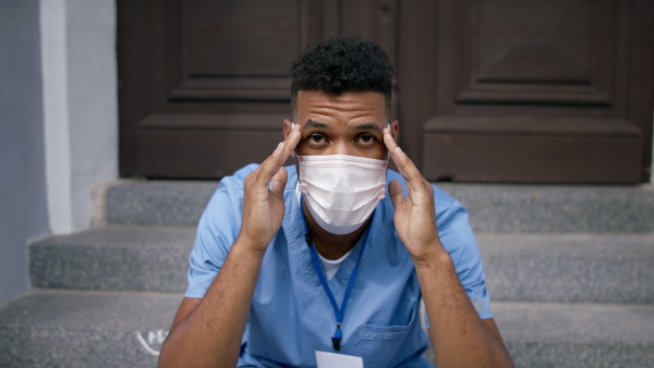 A frustrated male doctor sitting in front of the building with face mask and looking at camera.