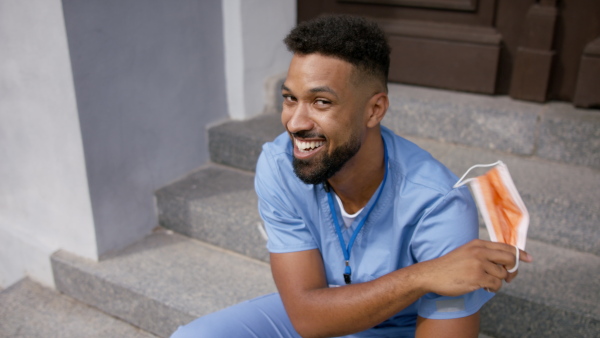 A happy male doctor sitting in front of the building taking off face mask and looking at camera.