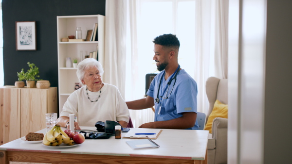 A happy healthcare worker or caregiver visiting senior woman indoors at home, measuring blood pressure.