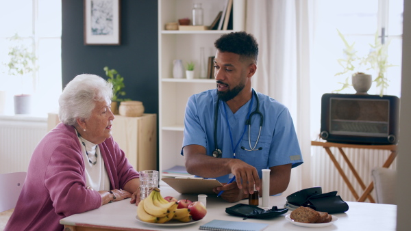 A happy healthcare worker or caregiver visiting senior woman indoors at home, talking and explaining dosage of pills.