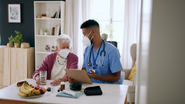 A happy healthcare worker or caregiver visiting senior woman indoors at home, talking and explaining, coronavirus concept.