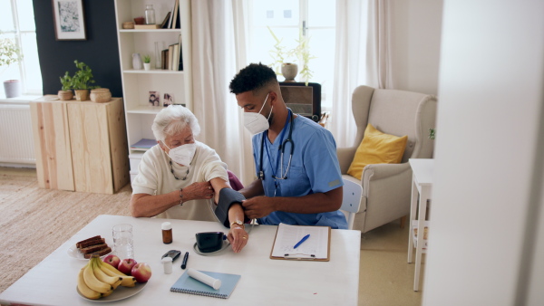 A happy healthcare worker or caregiver visiting senior woman indoors at home, measuring blood pressure, coronavirus concept.