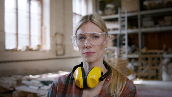 A female carpenter with goggles standing in workshop and looking at camera. Small business concept.