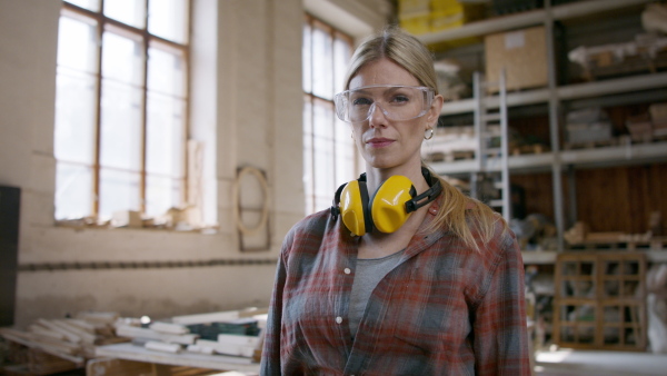 A female carpenter with goggles standing in workshop and looking at camera. Small business concept.