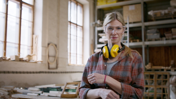 A female carpenter with goggles standing in workshop and looking at camera. Small business concept.