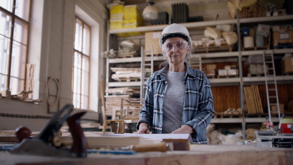 A senior female carpenter standing in workshop and looking at camera. Small business concept.