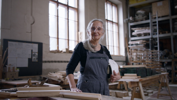 A senior female carpenter standing in workshop and looking at camera. Small business concept.