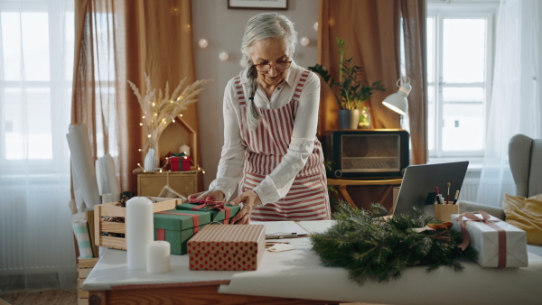 A senior woman packing Christmas presents and writing notes to list indoors, small business concept.