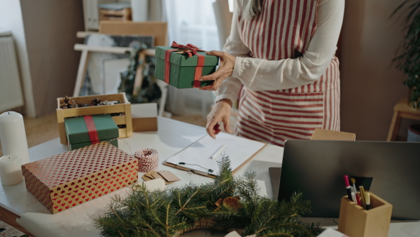 A senior woman packing Christmas presents and writing notes to list indoors, small business concept.