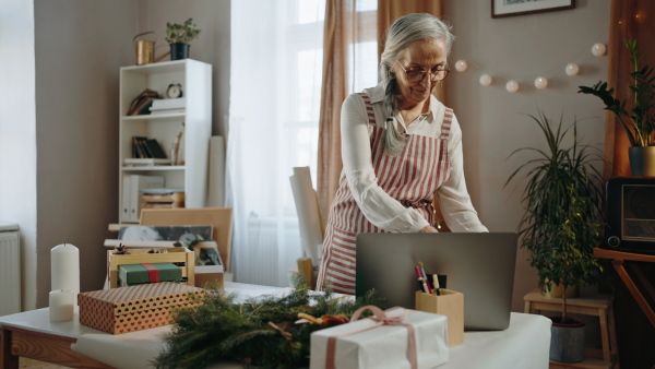 A senior woman packing Christmas presents and writing notes to list indoors, small business concept.