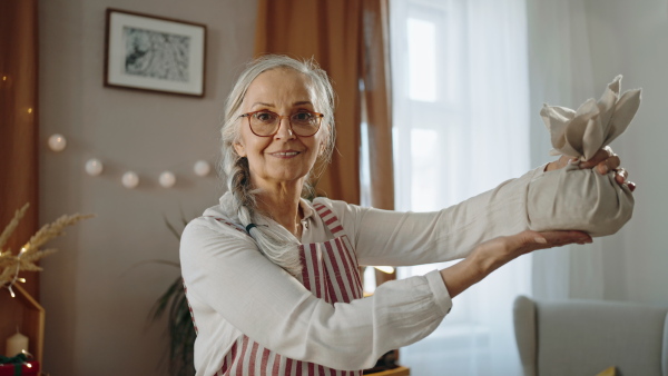 A senior woman looking at camera and packing Christmas present in natural materials indoors at home, environmental gift wrapping concept