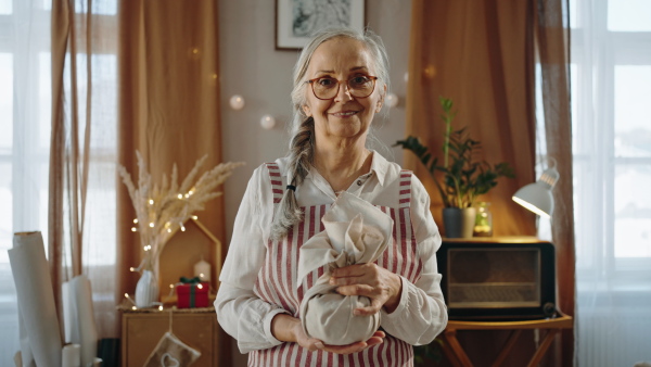 A senior woman looking at camera and packing Christmas present in natural materials indoors at home, environmental gift wrapping concept