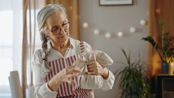 A senior woman packing Christmas present in natural materials indoors at home, environmental gift wrapping concept