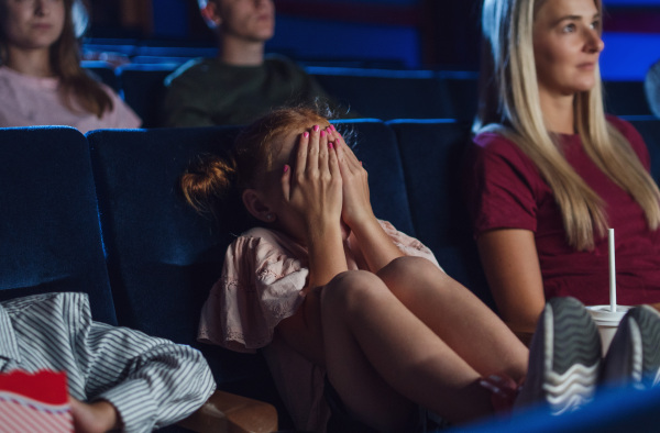 A worried small girl with mother covering eyes in the cinema while watching film.