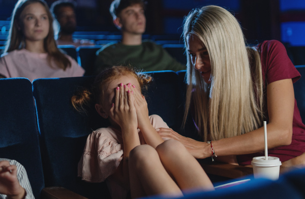 A worried small girl covering eyes in the cinema while watching film, mother comforting her.