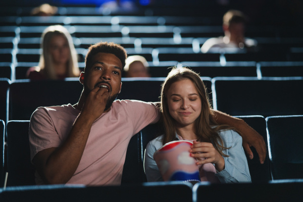 A front view of cheerful young couple with popcorn in the cinema, watching film.