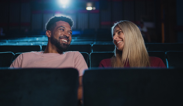 A front view of cheerful young couple in the cinema, watching film and talking.