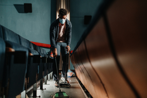 A young man cleaner sweeping floor in the cinema after the film, coronavirus concept.