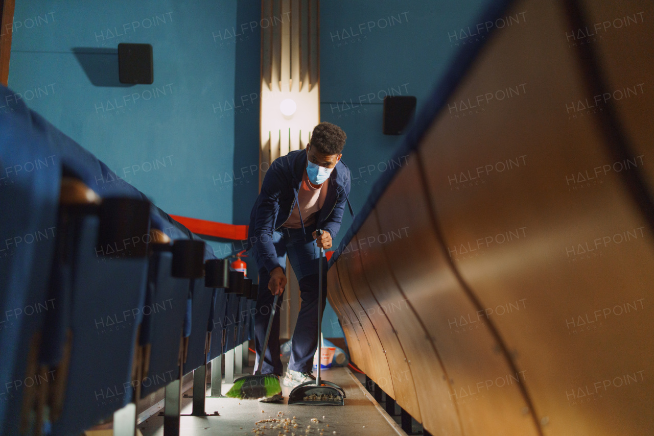 A young man cleaner sweeping floor in the cinema after the film, coronavirus concept.