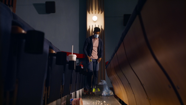 A young man cleaner sweeping floor in the cinema after the film, coronavirus concept.