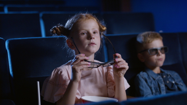 Happy small children with 3d glasses and popcorn in the cinema, watching film. Girl looking at camera.