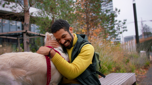 A happy young man sitting and embracing his dog during walk outdoors in city in autumn.