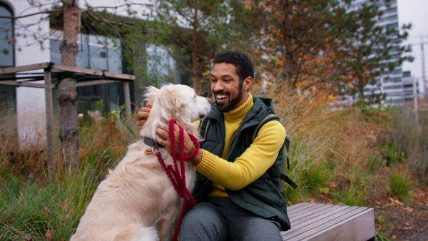 A happy young man sitting and stroking his dog during walk outdoors in city in autumn.