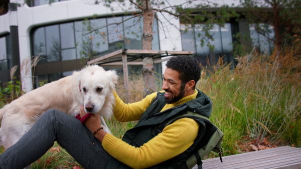 A happy young man sitting and stroking his dog during walk outdoors in city in autumn.