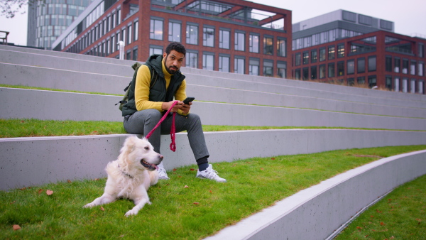 A happy young man sitting, resting and using smarthpone during dog walk outdoors in town, looking at camera.