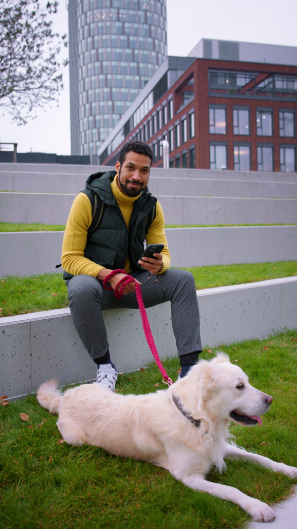 A vertical footage of happy young man sitting, resting and using smarthpone during dog walk outdoors in town, looking at camera.