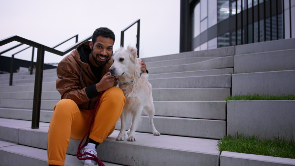 A happy young man sitting and embracing his dog and looking at camera outdoors in city in autumn.