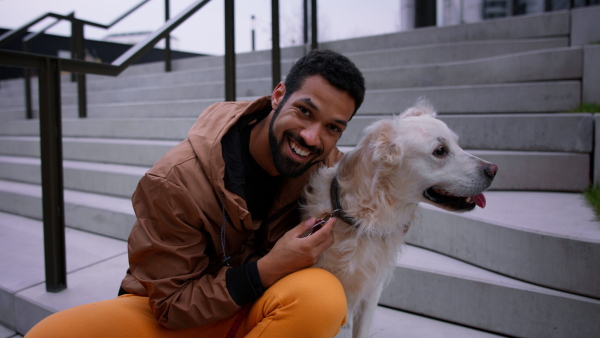 A happy young man sitting and embracing his dog and looking at camera outdoors in city in autumn.