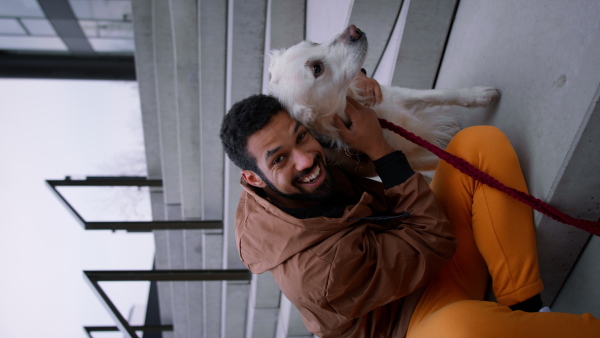 A vertical footage of happy young man sitting and embracing his dog and looking at camera outdoors in city in autumn.