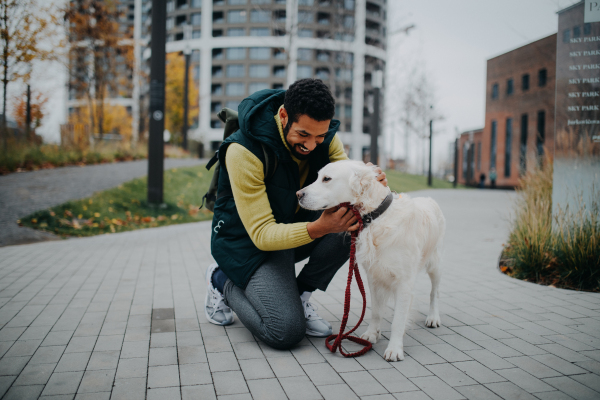 Happy young man stroking his dog outdoors in city park, during a cold autumn day.