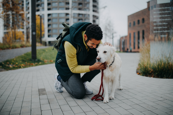 A happy young man squatting and stroking his dog during walk outdoors in city.