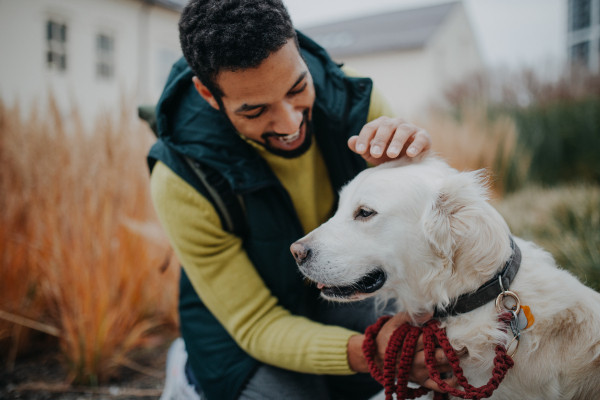 Happy young man stroking his dog outdoors in city park, during a cold autumn day.