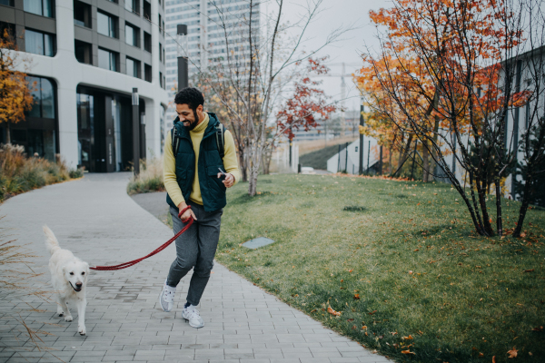 A happy young man walking his dog outdoors in city during autumn day.