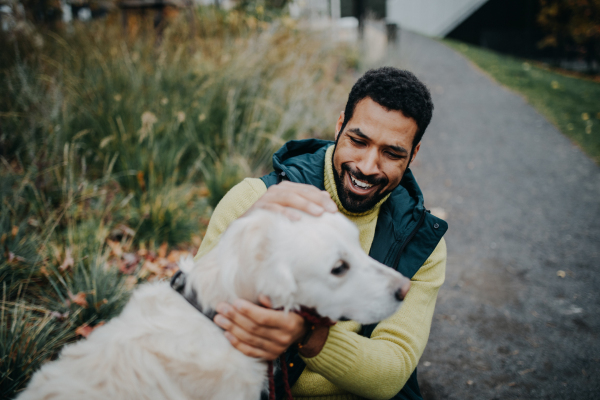 Happy young man stroking his dog outdoors in city park, during a cold autumn day.