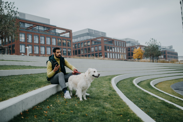 Happy young man resting and phoneing in urban city area during dog walk outdoors in cloudy autumn day.