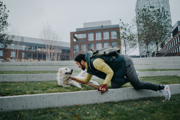 Happy young man training and cuddling with his dog outdoors in city park, during a cold autumn day.