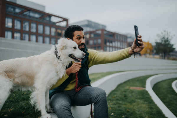 Happy young man resting and taking selfie with his dog outdoors in city park, during a cold autumn day.