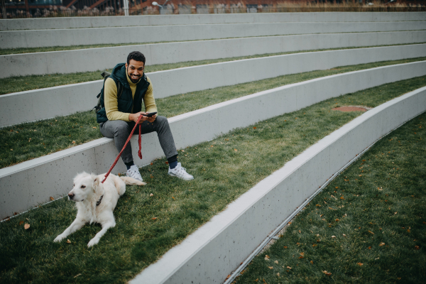 Happy young man resting and phoneing in urban city area during dog walk outdoors in cloudy autumn day.